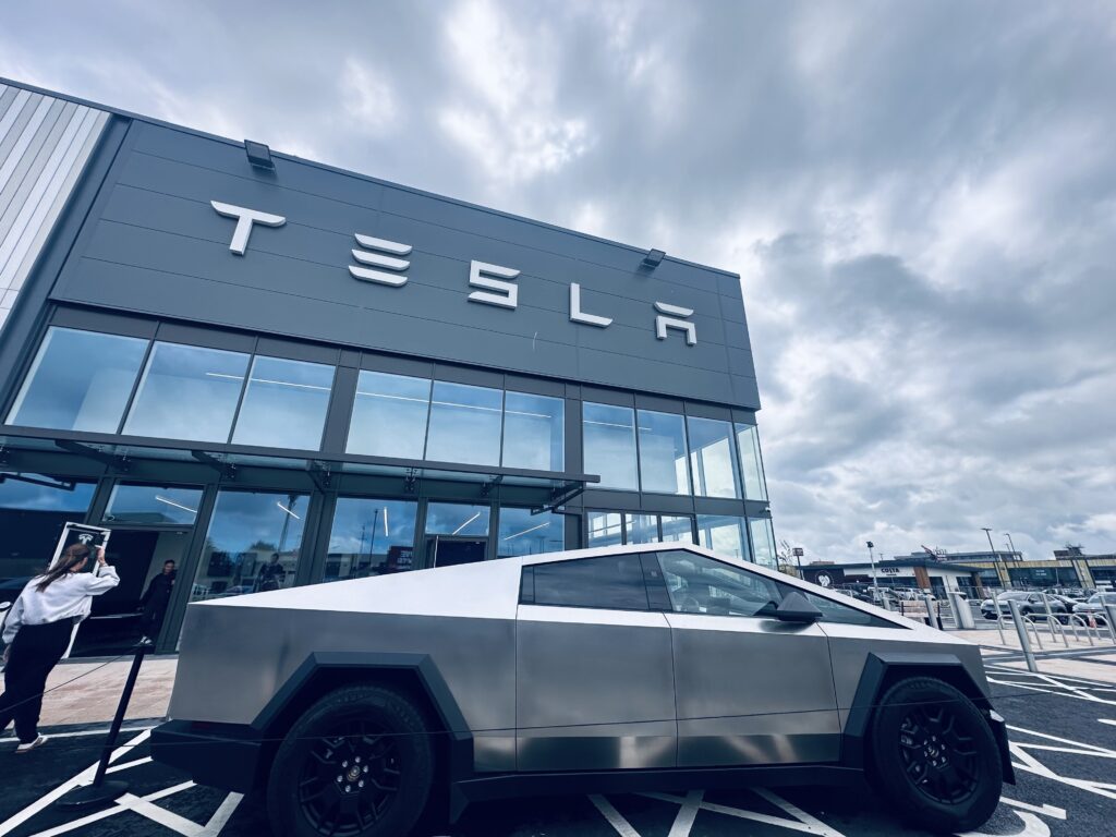 Tesla Cybertruck parked outside the new Tesla showroom at Liverpool Shopping Park, with modern glass-fronted architecture and Costa Coffee in the background.