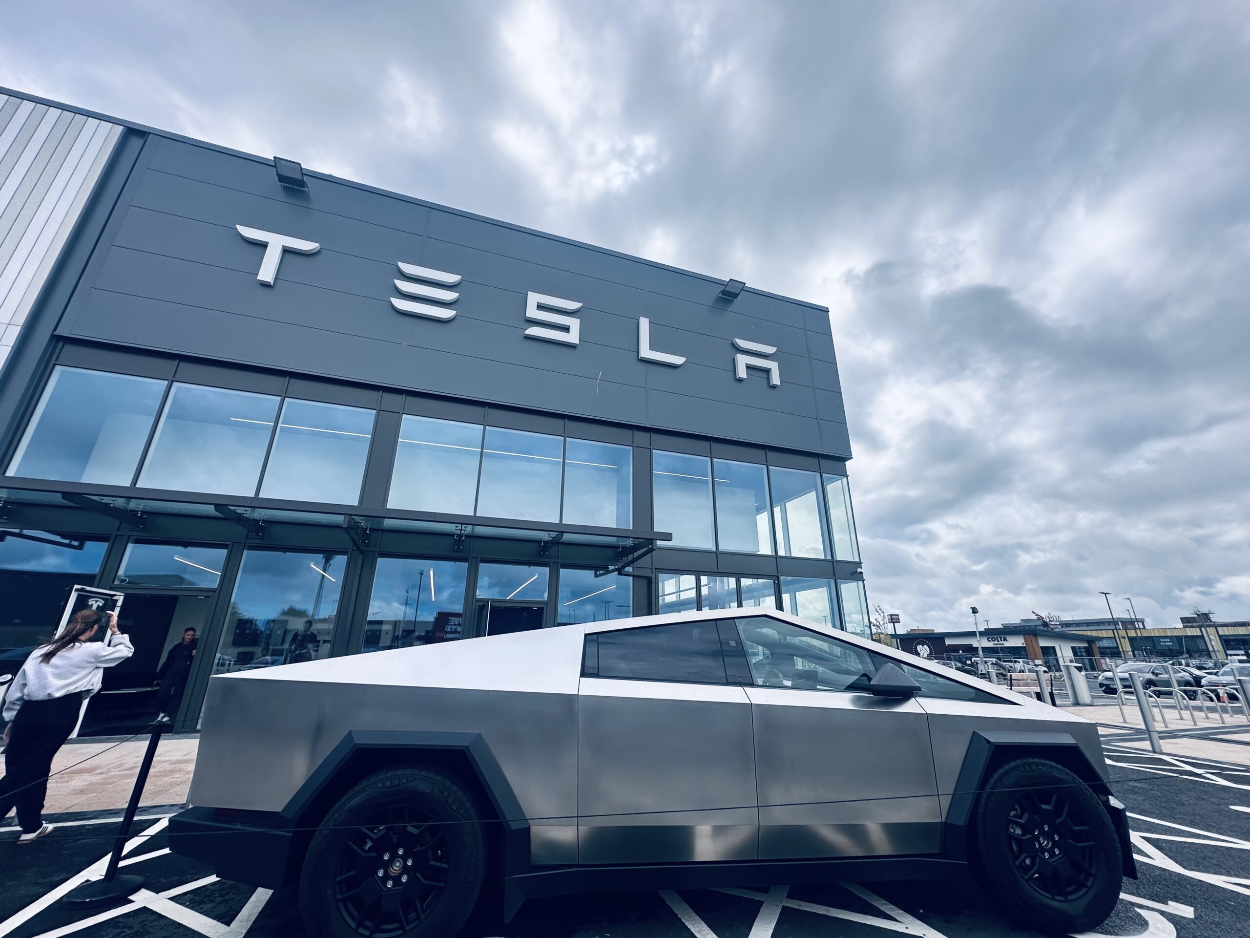 Tesla Cybertruck parked outside the new Tesla showroom at Liverpool Shopping Park, with modern glass-fronted architecture and Costa Coffee in the background.